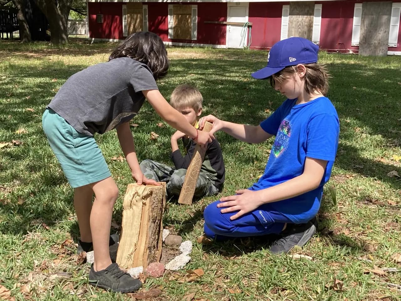 Learners practice outdoor skills on the grass beside a red barn, steadying a log on a stump.